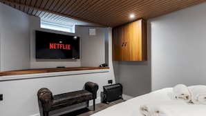 Un dormitorio moderno con techo de madera, un televisor grande mostrando 'NETFLIX', un banco, una maleta y una cama bien hecha con toallas.