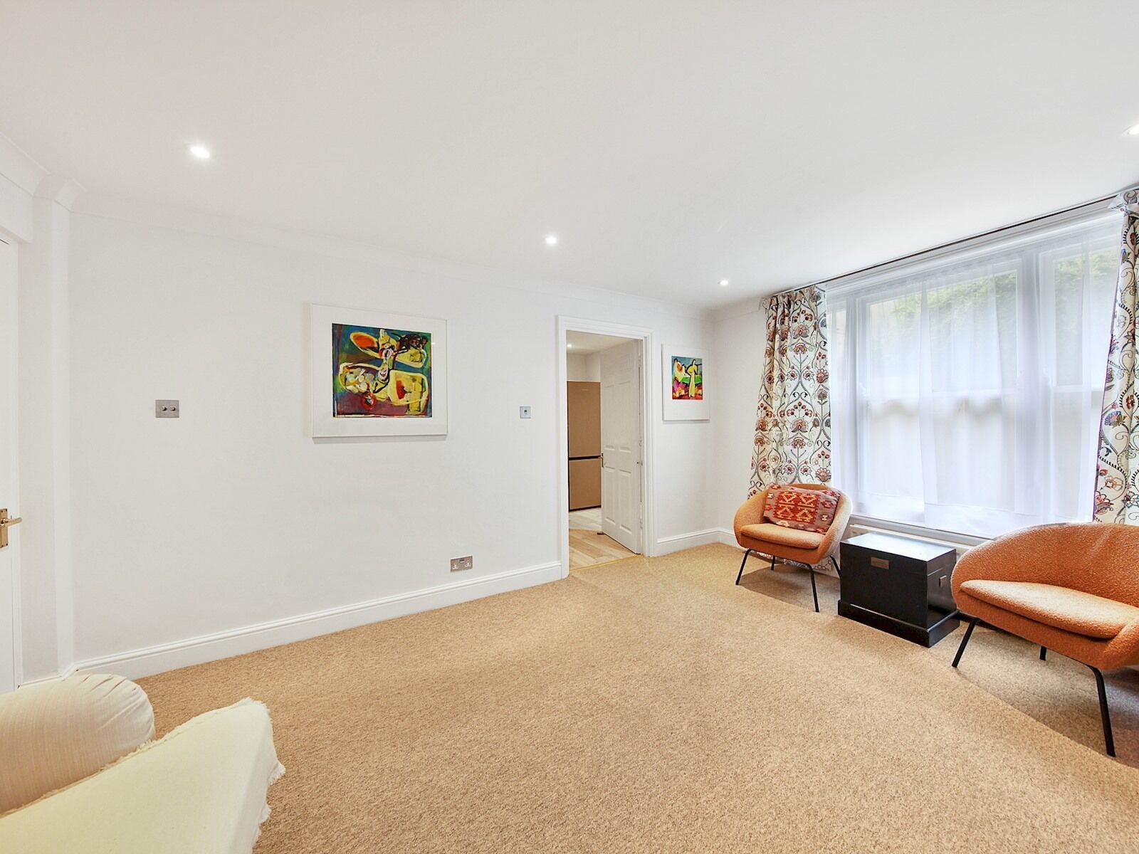 A spacious living room with a beige carpet, two armchairs, a sofa, and two framed artworks on the walls.