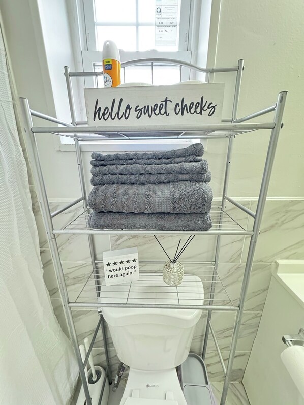A bathroom with a metal shelf holding folded towels, a toilet, and a sign that reads "hello sweet cheeks". - 1 Bedroom Urban Loft Suite near EWR, NYC, MetLife! (Newark)