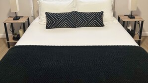 A bedroom with a large bed, two bedside tables with lamps, a window with curtains, and a black throw blanket. - 1 Bedroom Urban Loft Suite near EWR, NYC, MetLife! (Newark)
