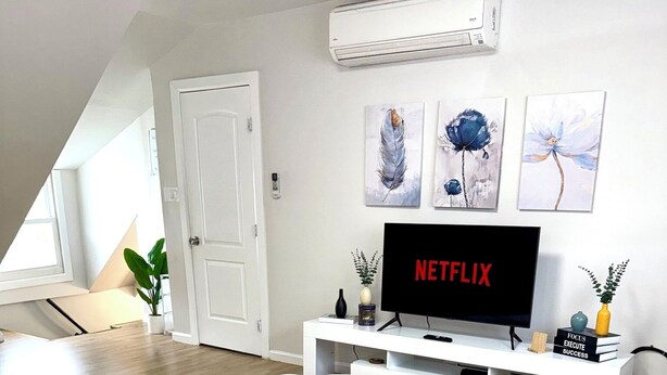 A modern living room with a white sofa, two white chairs, a white entertainment unit with a TV displaying 'NETFLIX', and a large window with a view of the sky. - 1 Bedroom Urban Loft Suite near EWR, NYC, MetLife! (Newark)