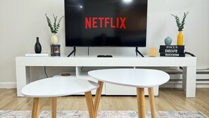 A modern living room with a flat-screen TV displaying 'NETFLIX', a white entertainment unit, two white round tables with wooden legs, a patterned rug, and three wall-mounted paintings. - 1 Bedroom Urban Loft Suite near EWR, NYC, MetLife! (Newark)