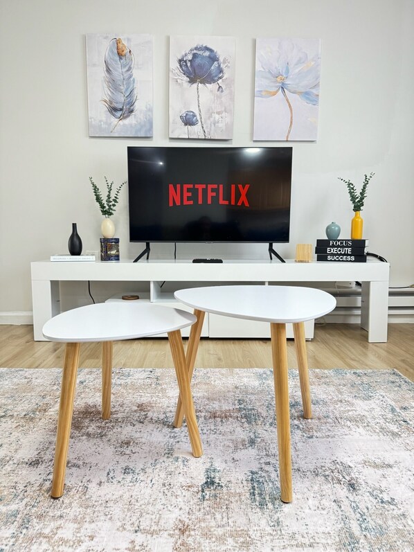 A modern living room with a flat-screen TV displaying 'NETFLIX', a white entertainment unit, two white round tables with wooden legs, a patterned rug, and three wall-mounted paintings.