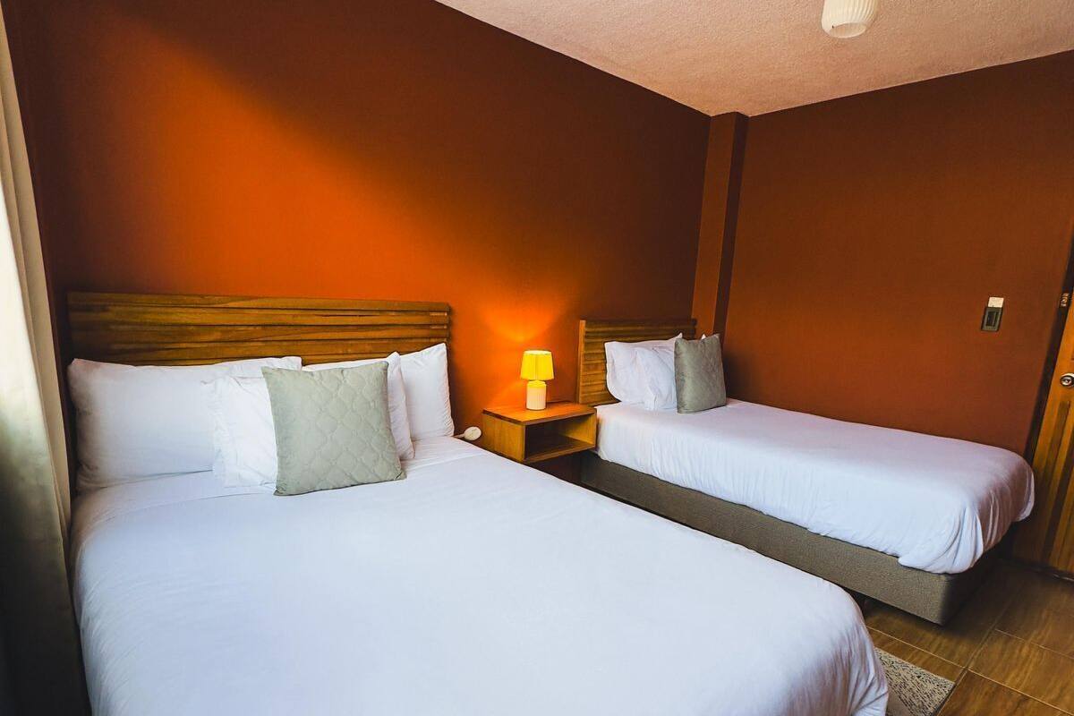 Une chambre d’hôtel avec deux lits, des têtes de lit en bois, du linge de lit blanc, une table de chevet avec une lampe et un luminaire au plafond.