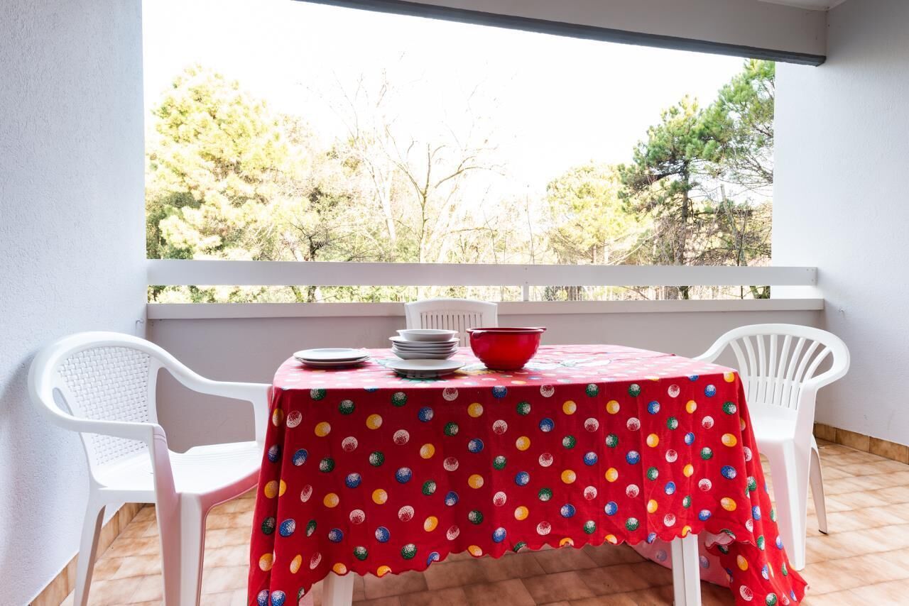 Un balcon avec une table recouverte d’une nappe rouge à pois, des chaises en plastique blanches et un bol rouge posé sur la table.