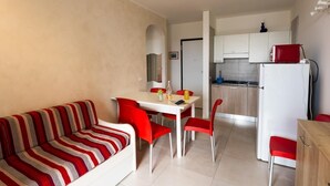 Un espacio habitable compacto con un sofá a rayas, una mesa de comedor con sillas rojas, una zona de cocina con armarios y un refrigerador, y un pequeño mueble de madera.