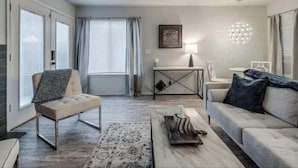 A modern living room with a sofa, a coffee table, and a side table. There is a window with curtains, a lamp, and a framed picture on the wall. - Cozy 1BR in Prime Location (Oklahoma City)