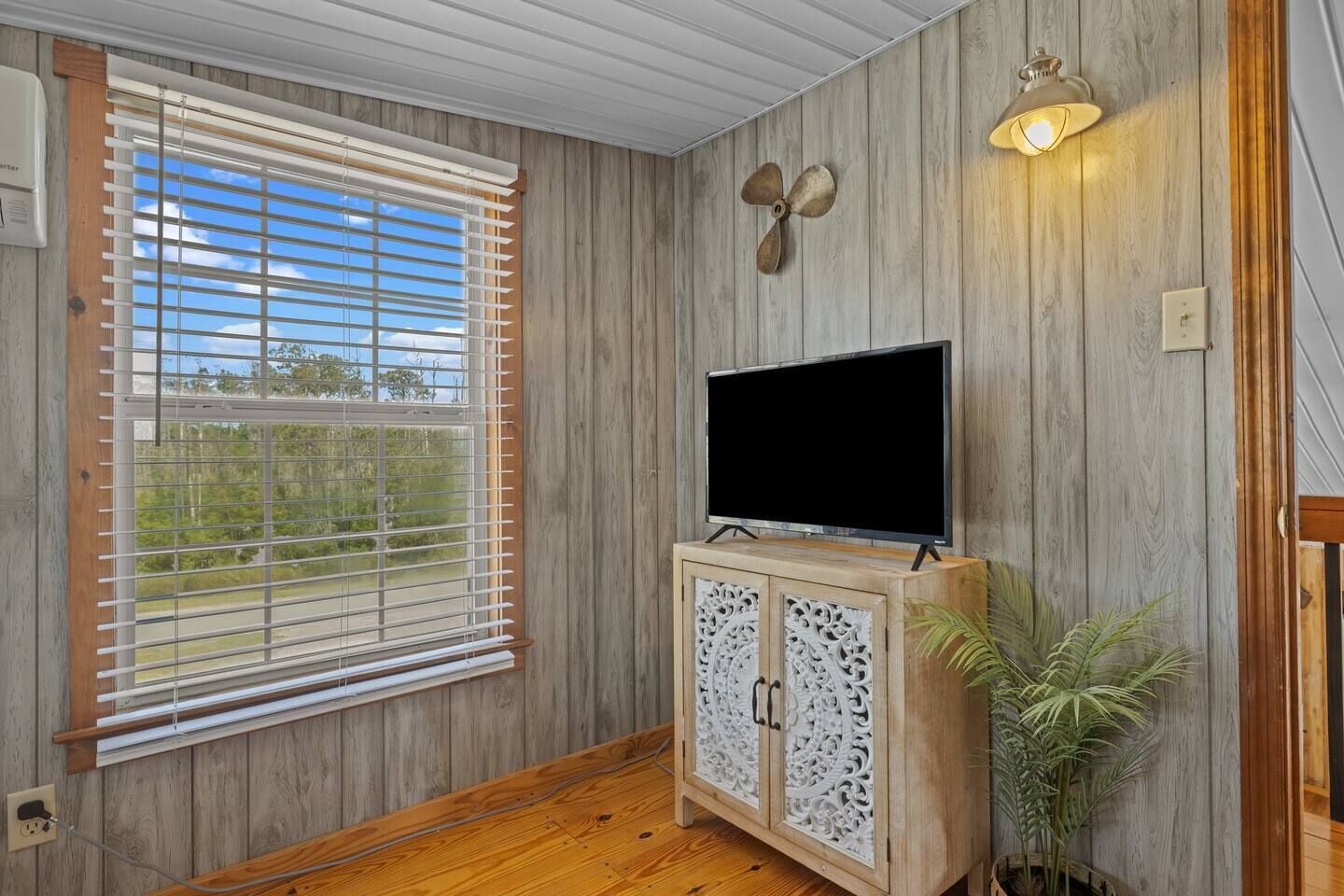 A room with a wooden paneled wall, a television on a cabinet, a ceiling fan, and a window with blinds.