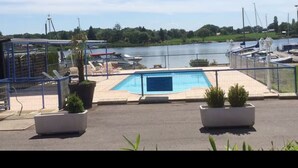 Zona de piscina con vista a un lago y barcos, rodeada por una cerca y plantas en macetas.