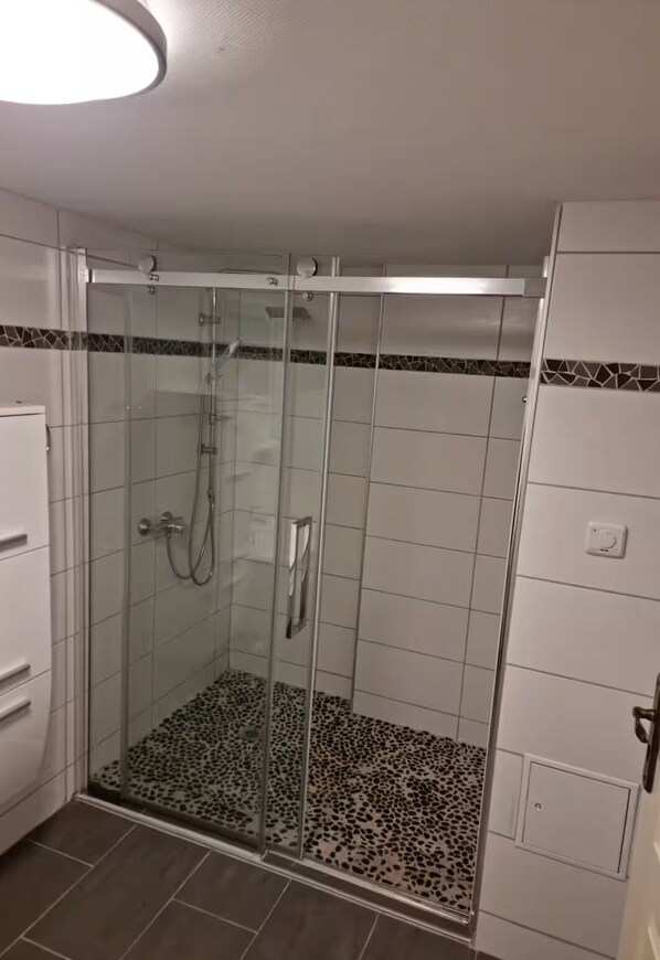 Ducha con cerramiento de vidrio, suelo de baldosas y un armario blanco a la izquierda.