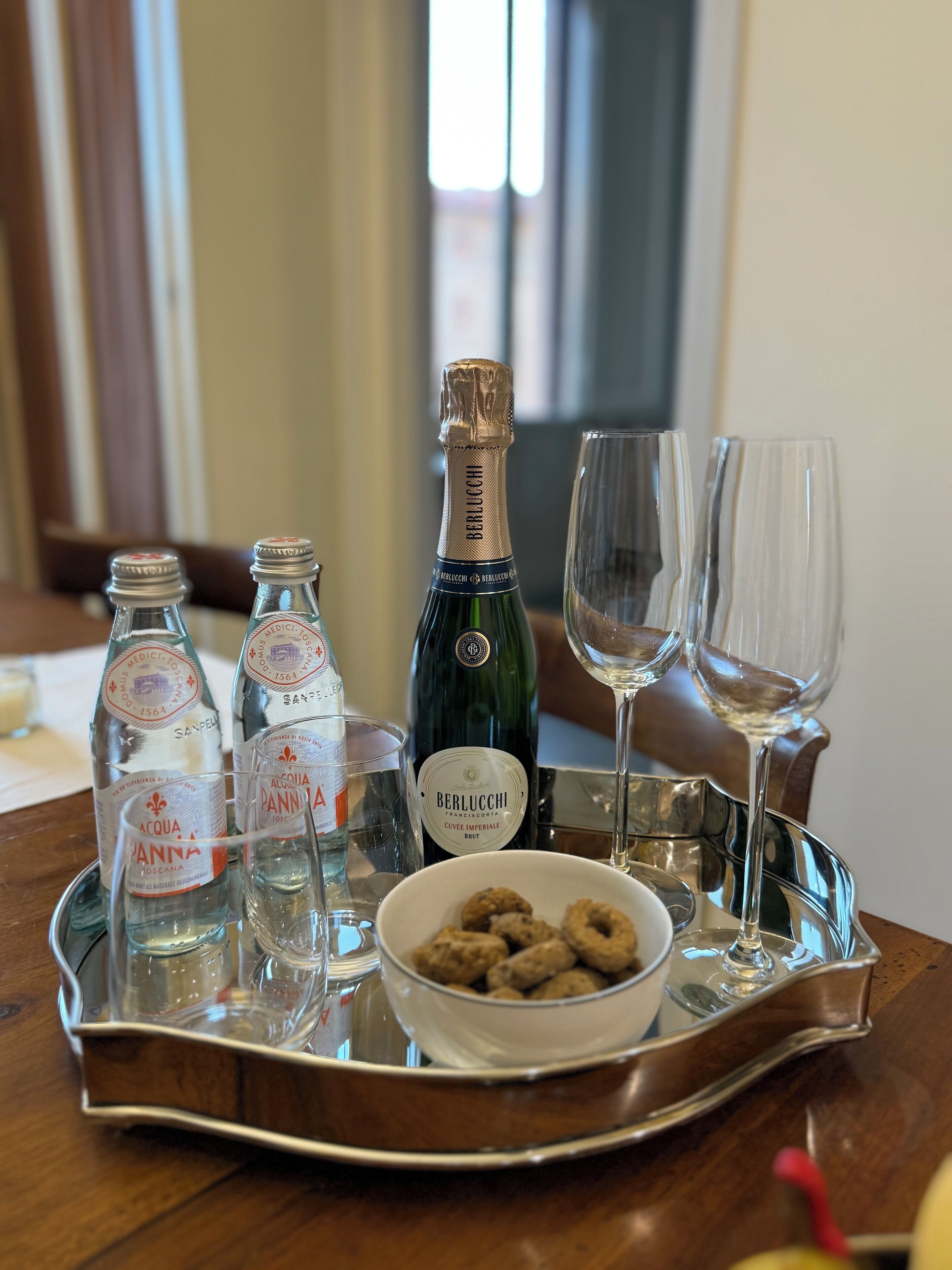Una bandeja con botellas de agua, una botella de champán, dos copas y un bol de aperitivos sobre una mesa de madera.