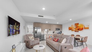 Una sala de estar moderna con un sofá, un sillón y una mesa de centro. Se ve una cocina al fondo, con un refrigerador y un microondas.