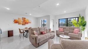 Una sala de estar moderna con un sofá, una mesa de comedor y sillas. Hay ventanas grandes y una planta.