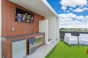 Una cocina exterior moderna con refrigerador incorporado, pileta y televisor de pantalla plana montado en una pared de madera. En primer plano, sillas de mimbre blancas y una mesa pequeña, y al fondo, una vista del horizonte de la ciudad.