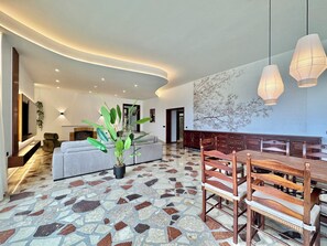 A modern living room with a grey sofa, a dining area with wooden chairs and a table, a large potted plant, and a wall-mounted TV. - GOLF SEA&POOL CACTUS VILLA (Buenavista del Norte)