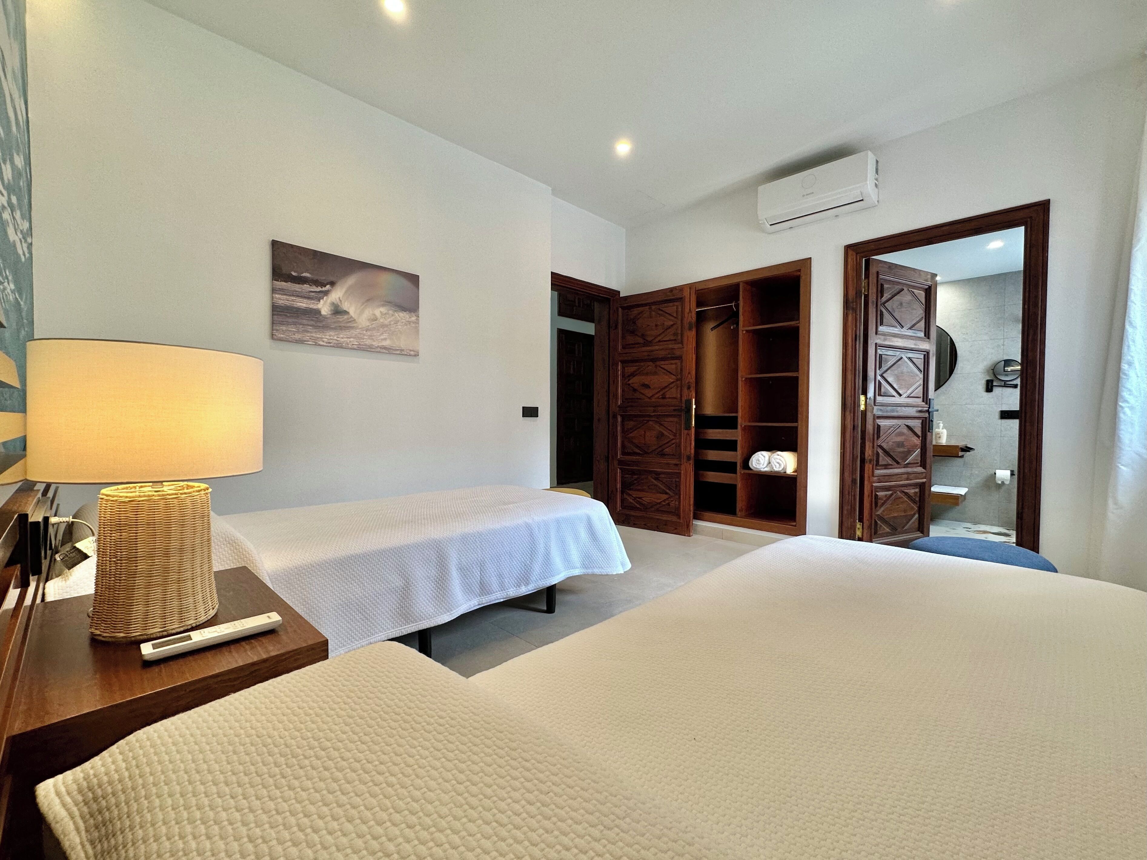 Habitación de hotel con dos camas, una mesita de noche con lámpara, un armario y un baño visible a través de una puerta abierta.