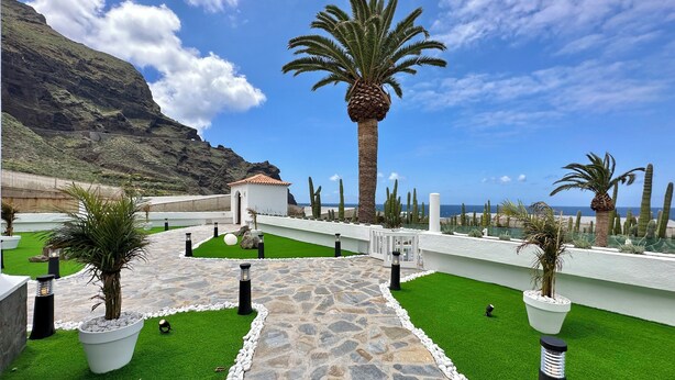 A landscaped pathway leading to a white building with a red-tiled roof, surrounded by cacti and palm trees, with a mountainous backdrop. - GOLF SEA&POOL CACTUS VILLA (Buenavista del Norte)
