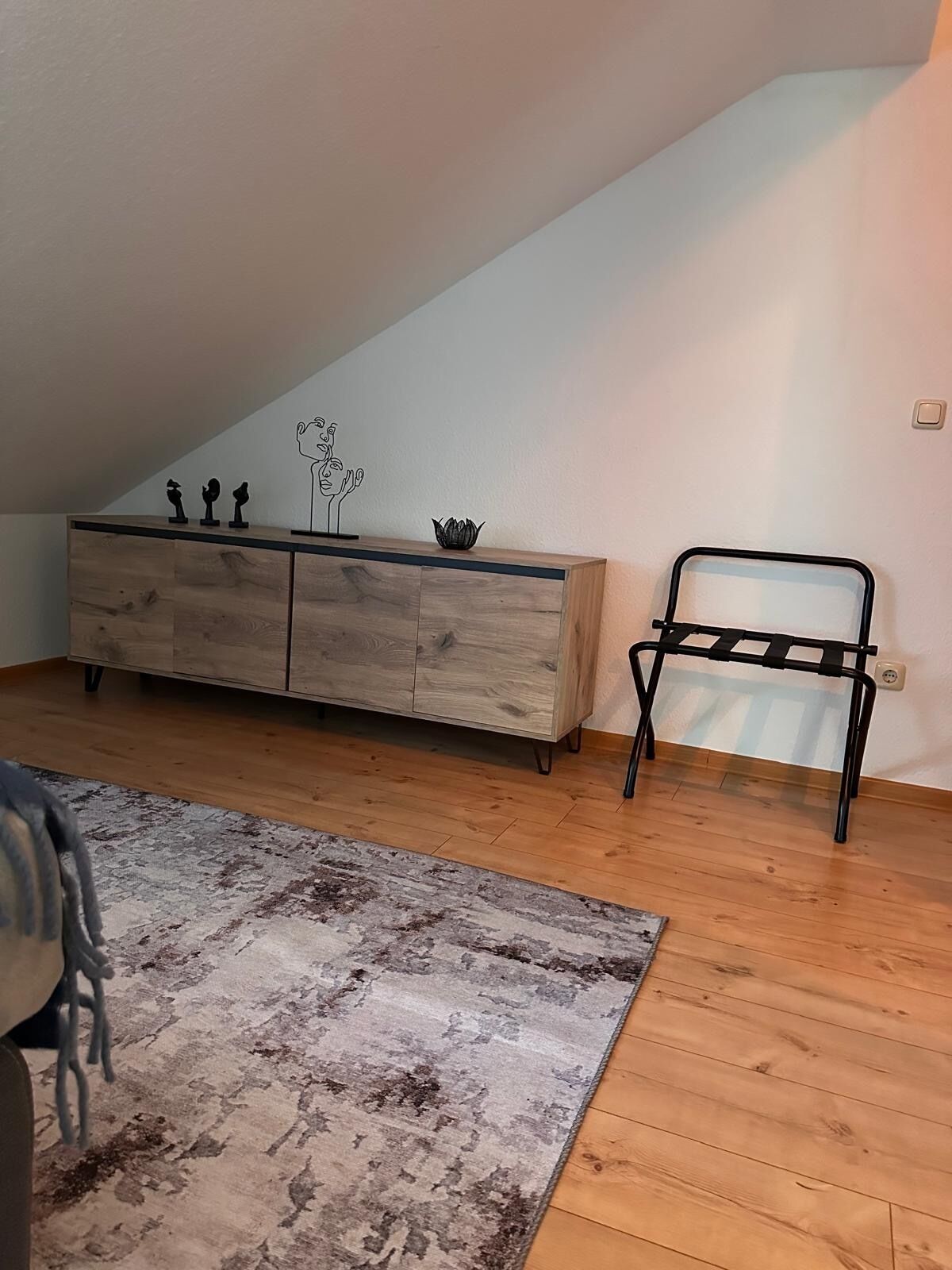 Une pièce avec un plafond en pente, un plancher en bois, un tapis à motifs gris, un meuble en bois orné d’objets décoratifs et un banc en métal noir.