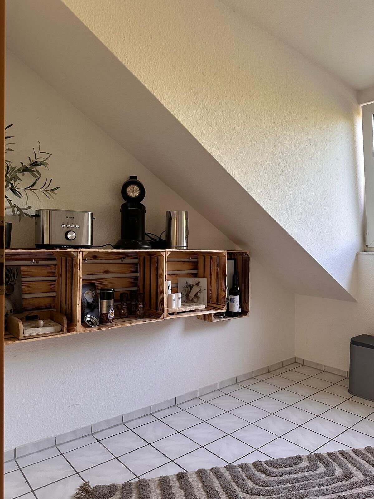 Een keuken met een houten wandplank waarop keukenapparatuur en voorwerpen staan, een betegelde vloer en een trap die naar een hoger niveau leidt.