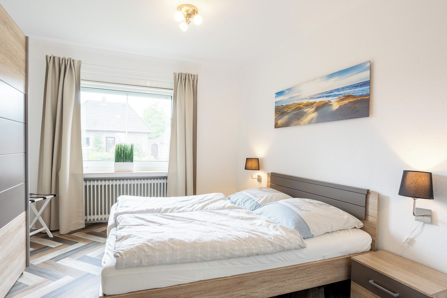 A bedroom with a bed, two bedside lamps, a window with curtains, a radiator, and a painting on the wall.