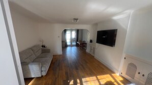 A spacious living area with a wooden floor, a grey sofa, a flat-screen TV mounted on the wall, and a dining area in the background. - Charming Houghton Regis House With 4 Bedrooms (Dunstable)