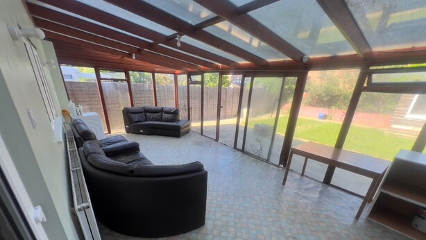 A covered patio area with a black sectional sofa, a wooden table, and a glass door leading to a backyard. - Charming Houghton Regis House With 4 Bedrooms (Dunstable)