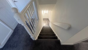 A staircase with white railings and a carpeted floor leading to a room with a door and a light fixture. - Charming Houghton Regis House With 4 Bedrooms (Dunstable)