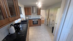 A compact kitchen with wooden cabinets, a black countertop, a built-in oven, and a white tiled floor. - Charming Houghton Regis House With 4 Bedrooms (Dunstable)