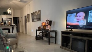 A modern living room with a flat-screen TV, a dining area with a glass table, and a kitchen in the background.