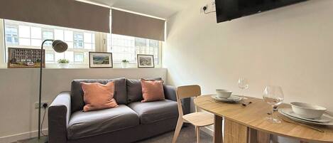 A modern living room with a grey sofa, a wooden dining table, and a flat-screen TV mounted on the wall.