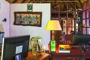 A room with a desk, computer, lamp, framed artwork, and a shelf with decorative items. - Bushfront Lodge (Livingstone)