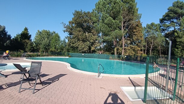 A swimming pool with a curved edge, surrounded by a brick patio and green fencing. - House in vacation residence 4 persons swimming pool, tennis, petanque, sneakers (Saint-Julien-en-Born)