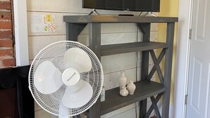 A room with a TV on a wooden TV stand, a white standing fan, and a fireplace.