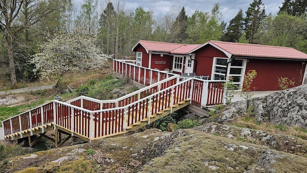 A red cabin with a wooden staircase leading to a deck, surrounded by trees and rocky terrain.