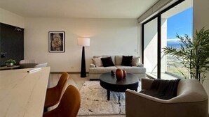 Una sala moderna con sofá, mesa de centro y sillas, un comedor con mesa y sillas, y grandes ventanas que ofrecen una vista.