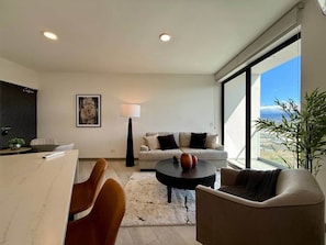 A modern living room with a sofa, coffee table, and chairs, a dining area with a table and chairs, and large windows offering a view.