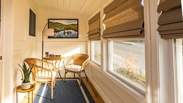 A room with a wooden floor, two wicker chairs, a blue rug, a small side table with a plant, and a painting on the wall. - Lake George Luxury Cabin (Alcove Studio) (Lake George)