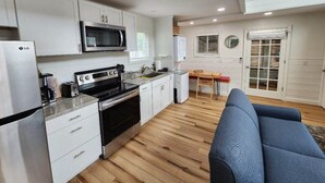 A compact kitchen with white cabinets, a microwave, a stove, and a refrigerator. There's a dining area with a table and chairs, and a blue sofa in the living space. - Lake George Luxury Cabin (Alcove Studio) (Lake George)