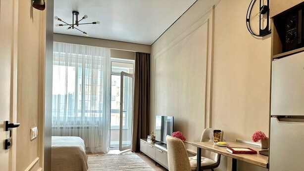 A modern hotel room with a bed, a desk with a chair, a television, and a refrigerator.