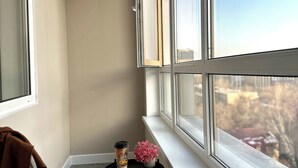 A corner room with a window, a chair, a small table with a cup and flowers, and a view of the city.