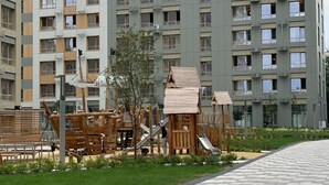 A playground area with wooden play structures and a small slide, surrounded by a paved walkway and greenery.