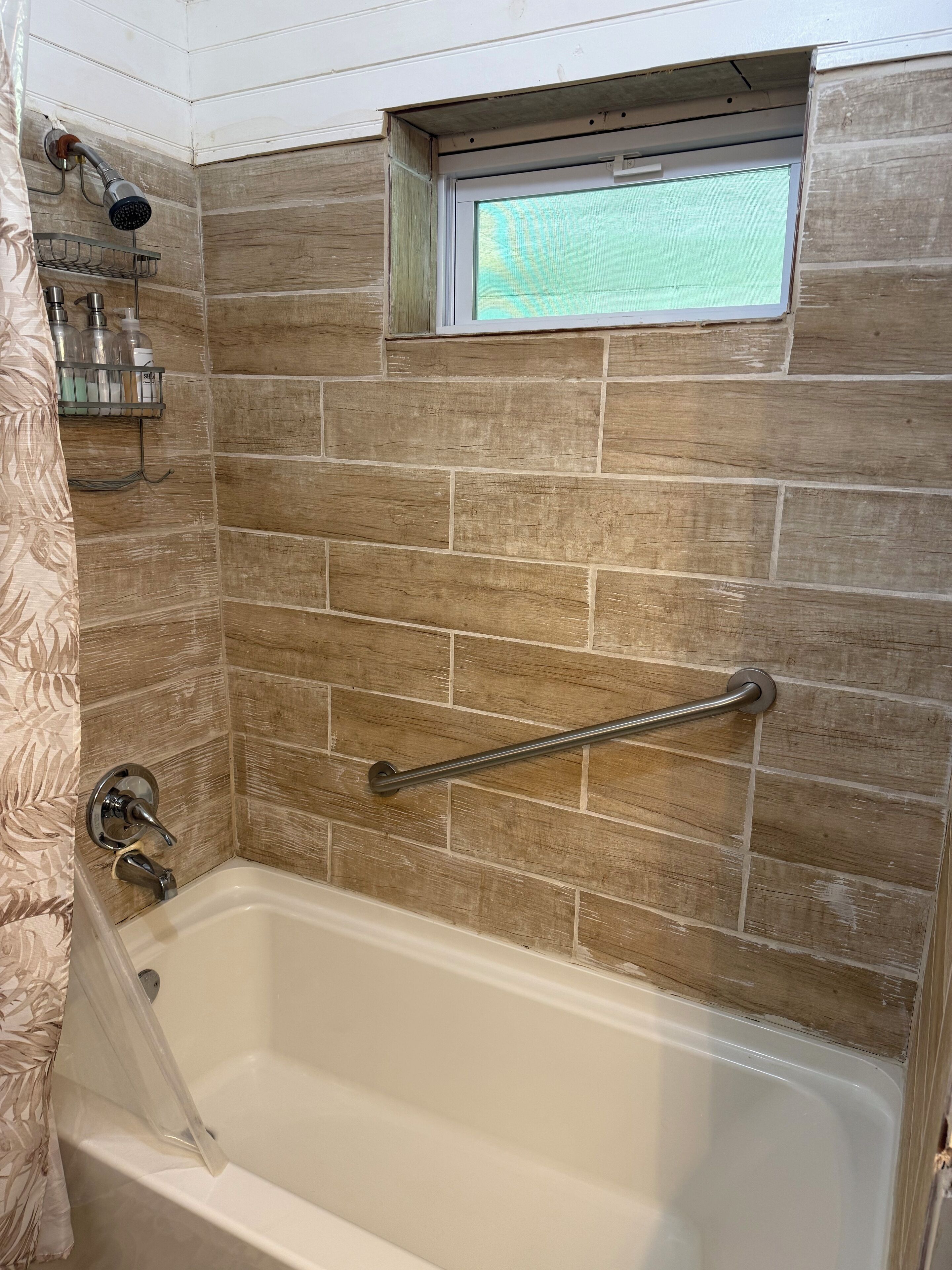 A bathroom with a bathtub, a showerhead, a window, and a handrail.