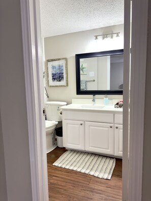 A bathroom with a white sink, mirror, and toilet. There is a framed picture on the wall and a small trash can beside the toilet.