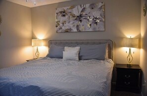 A bedroom with a large bed, two bedside tables, a wall-mounted lamp, and a floral artwork. - I207 - Apt|Patio|10mi to Convention Ctr|Sleeps 2 (Irving)