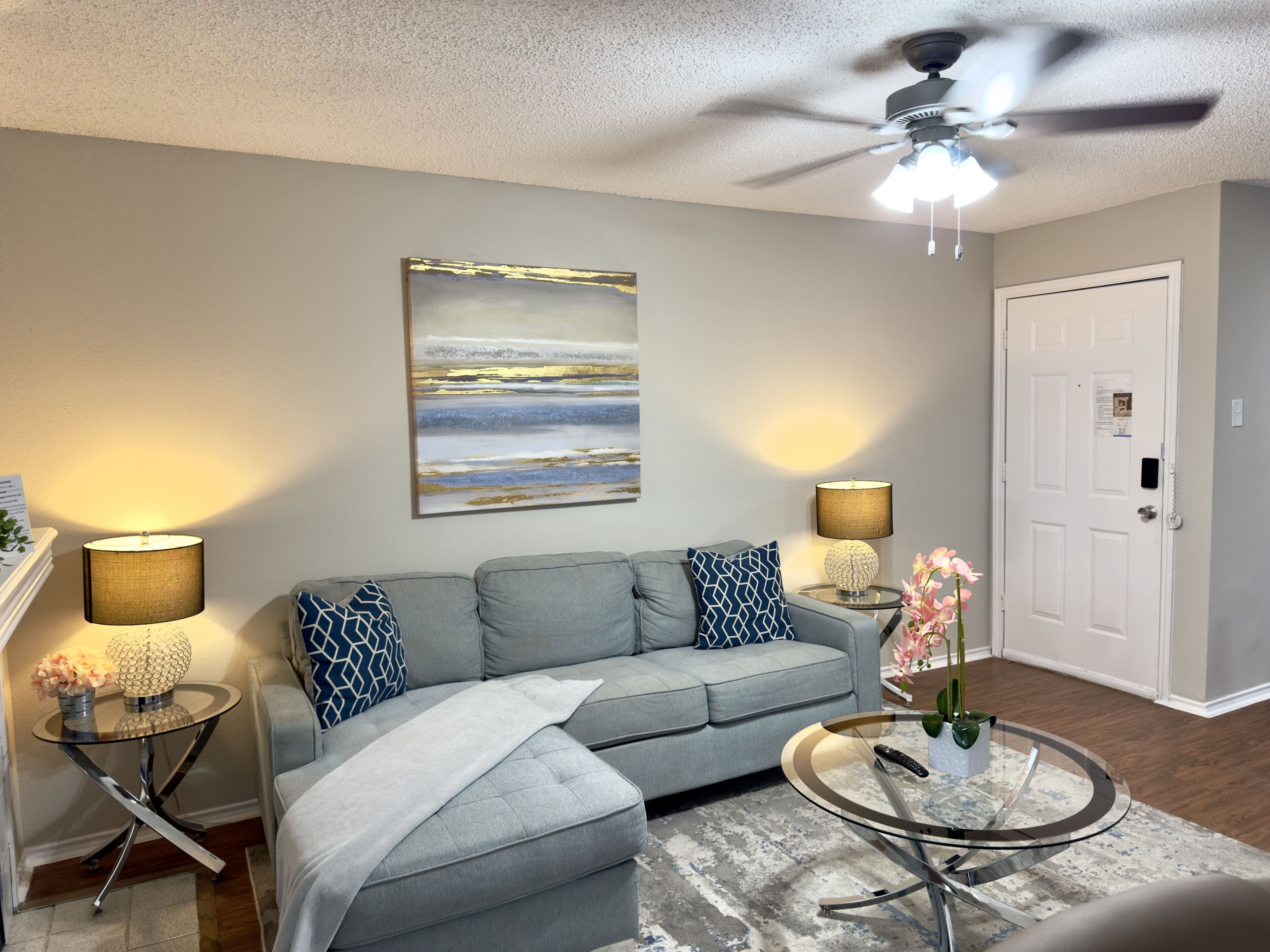 A living room with a grey sofa, two side tables with lamps, a ceiling fan, a door, and a large abstract painting on the wall.
