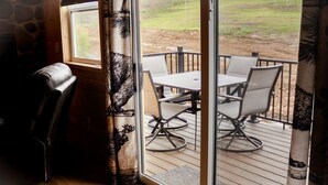 A room with a wooden floor, a glass door leading to an outdoor deck with a table and chairs, and a curtain with a nature design. - Entire Cabin in Linn, MO with all the extras (Linn)