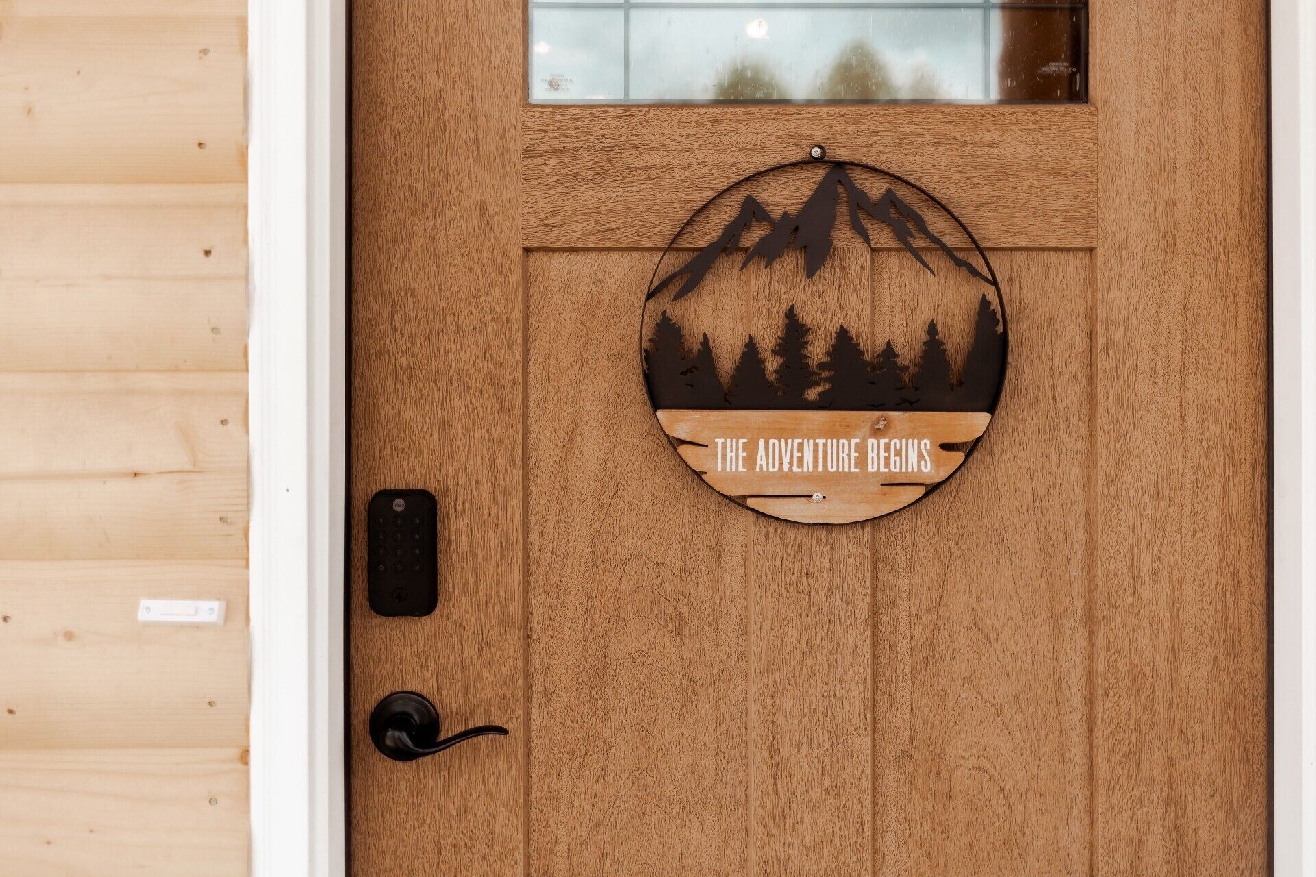 Una puerta de madera con un letrero circular que dice "THE ADVENTURE BEGINS" y presenta la silueta de una montaña y un bosque.