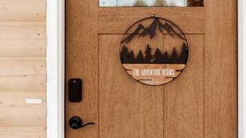 Una puerta de madera con un letrero circular que dice "THE ADVENTURE BEGINS" y presenta la silueta de una montaña y un bosque.