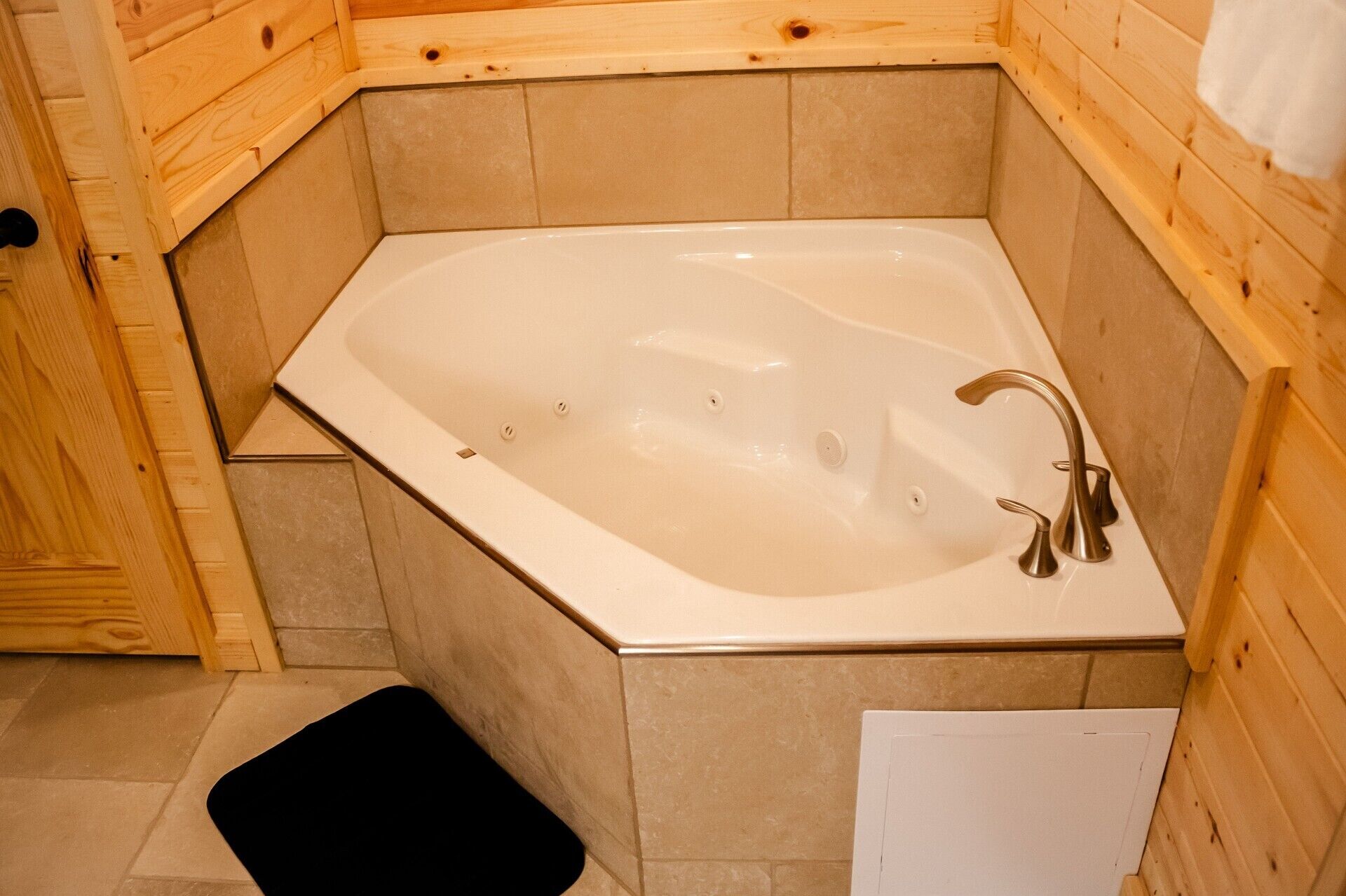 A corner bathtub with a wooden surround, a black bath mat, and a white towel on the wall.