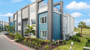 A modern multi-story building with a blue and gray facade, a covered outdoor area, and a landscaped garden with palm trees. - Golf View Penthouse w/ Rooftop Terrace Near Disney (Kissimmee)